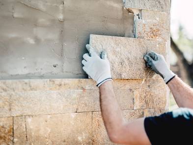 Fassadenkonstruktion, Hände eines Handwerkers, der gerade eine Steinplatte an eine Hauswand mauert, Foto: Bogdanhoda / iStock