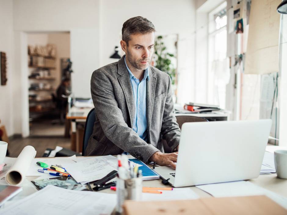 HOAI Leistungsphasen, Architekt an seinem Schreibtisch mit Unterlagen, Plänen und Laptop, Foto: Geber86 / iStock